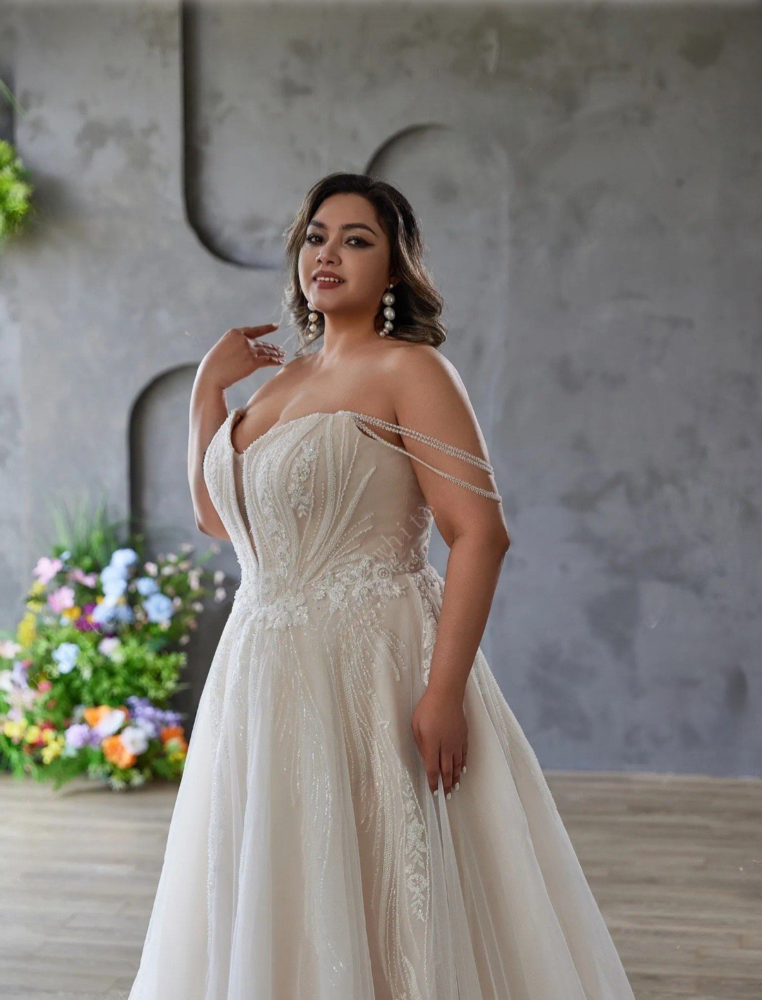 Woman in a white lace wedding dress standing in front of a gray wall with decorative elements.