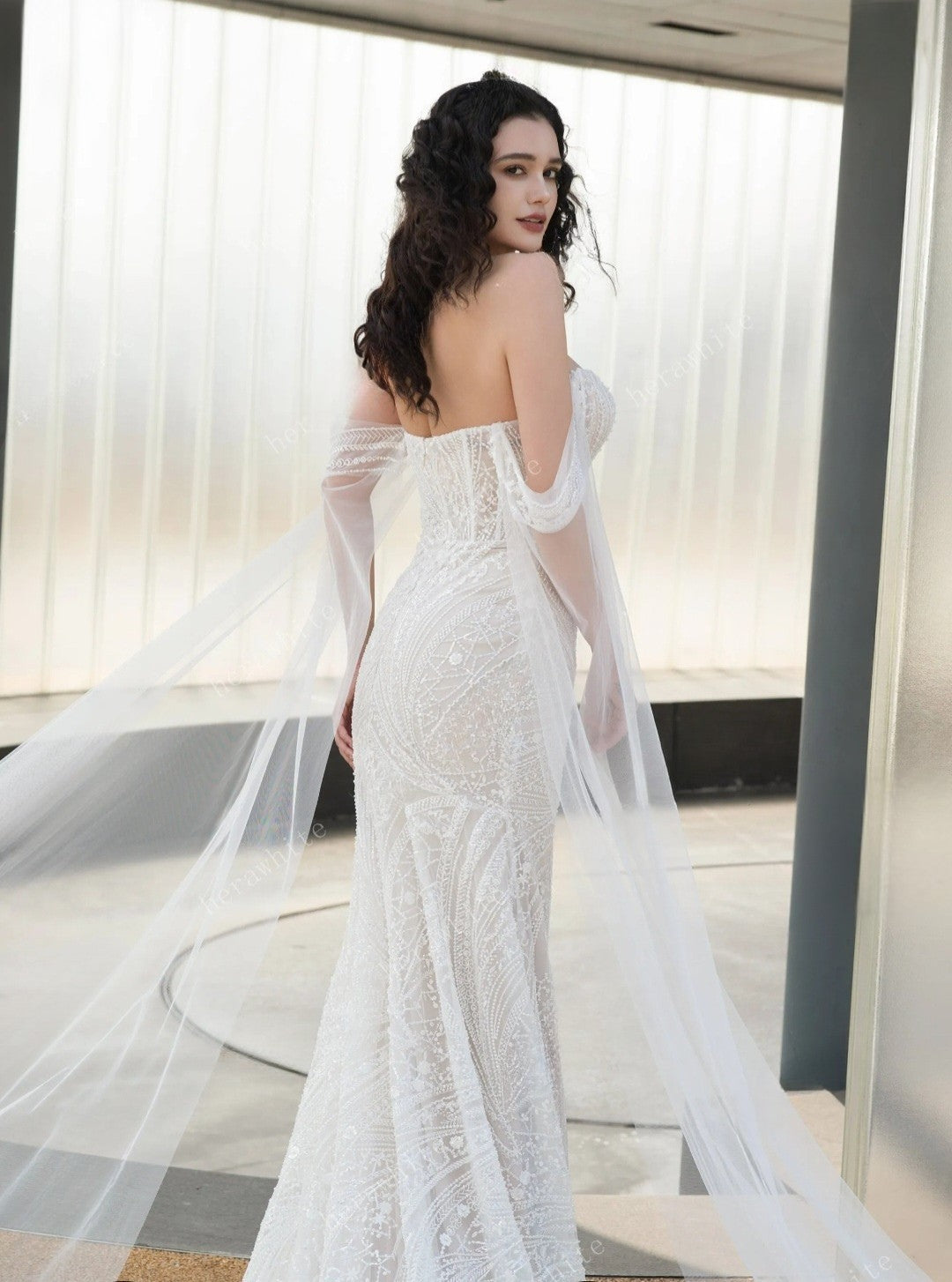 Woman in a white wedding dress with a long train standing indoors.