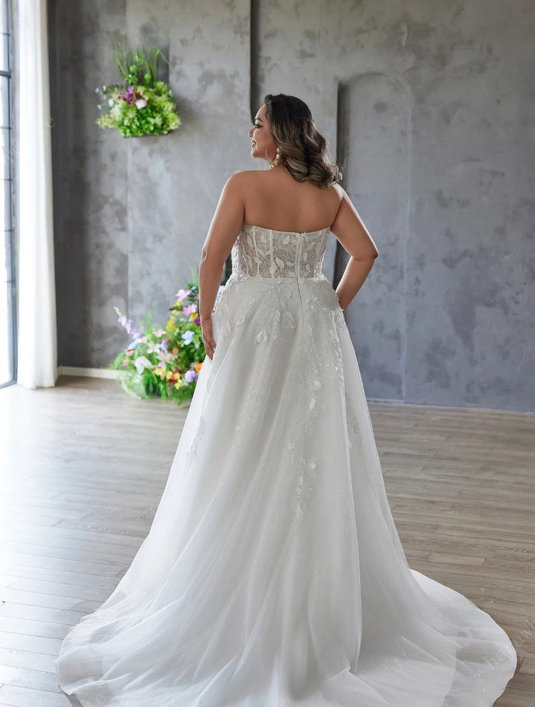Woman in a white wedding dress standing in a room with decorative flowers and a textured wall.