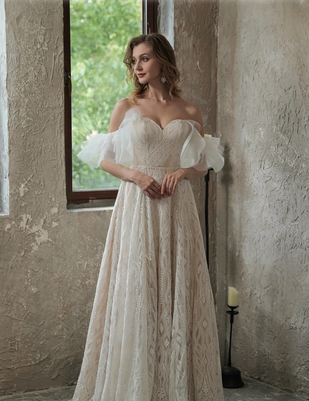 Woman in a white lace wedding dress standing in a room with a window and candle.