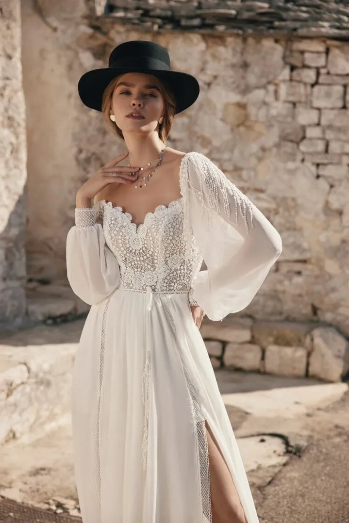 Woman wearing a white lace dress with long sleeves and a black hat in front of a stone wall.