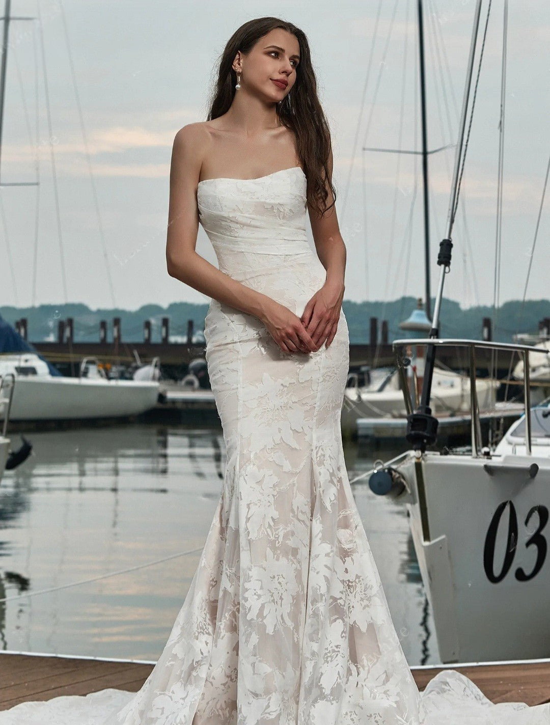Woman in a white strapless lace dress standing by a marina with boats in the background.