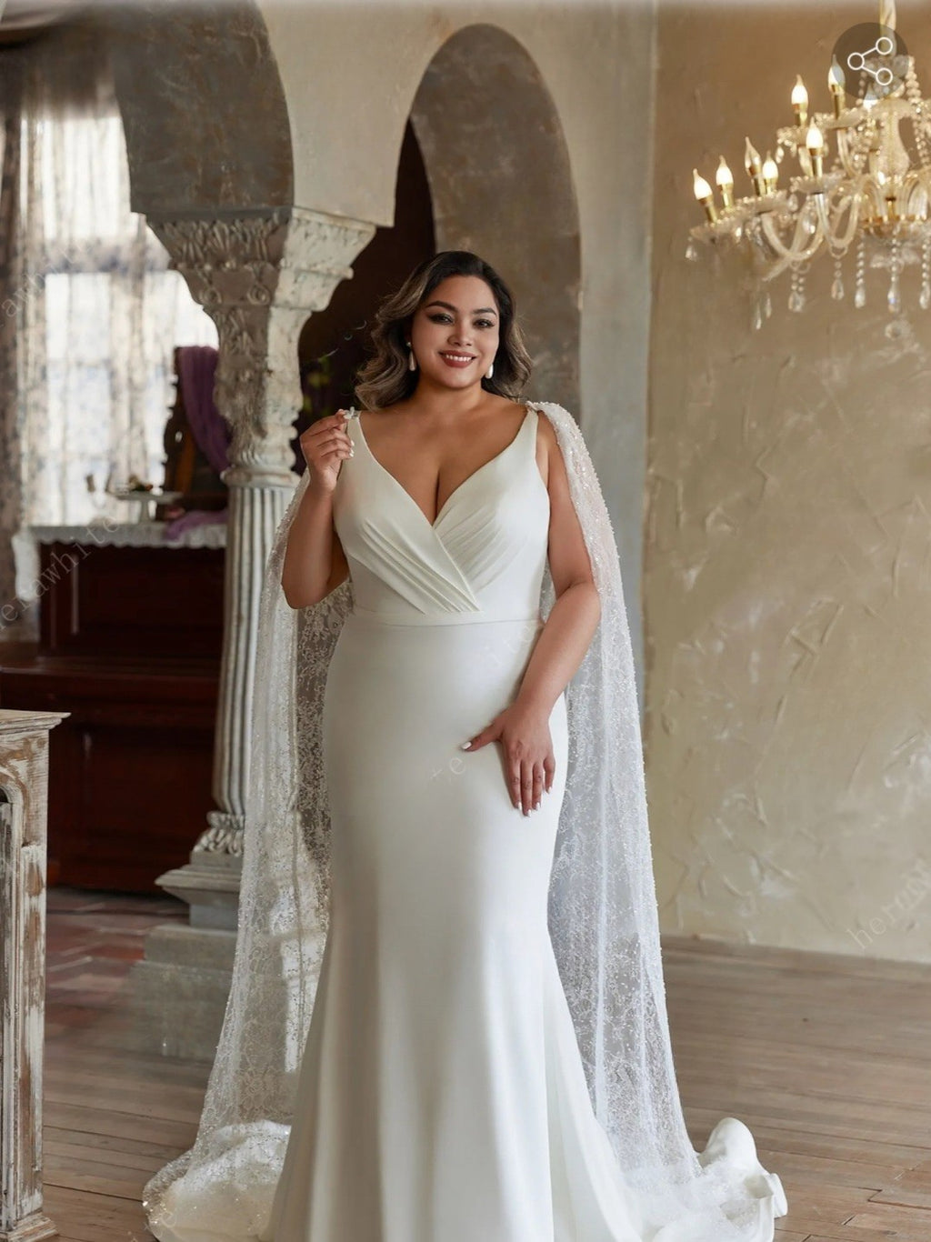 Woman in a white wedding dress with a veil standing in an elegant room with a chandelier.