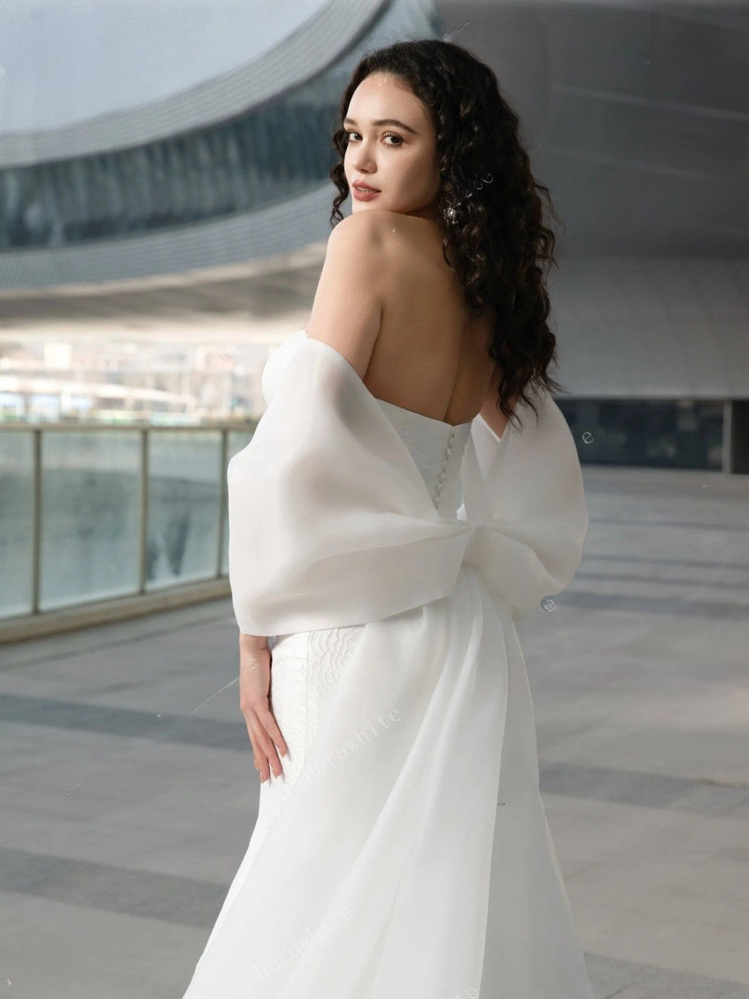 Woman in a white off-shoulder dress standing in an indoor setting with a modern architectural background.