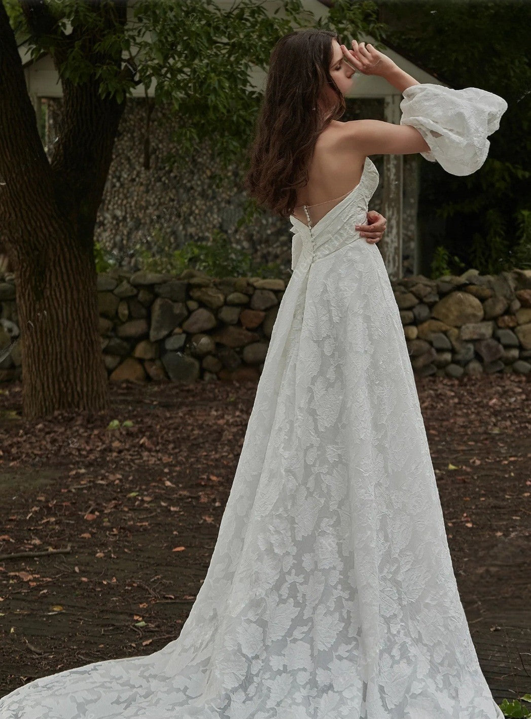 Woman in a lace wedding dress standing outdoors with trees and stone wall in the background