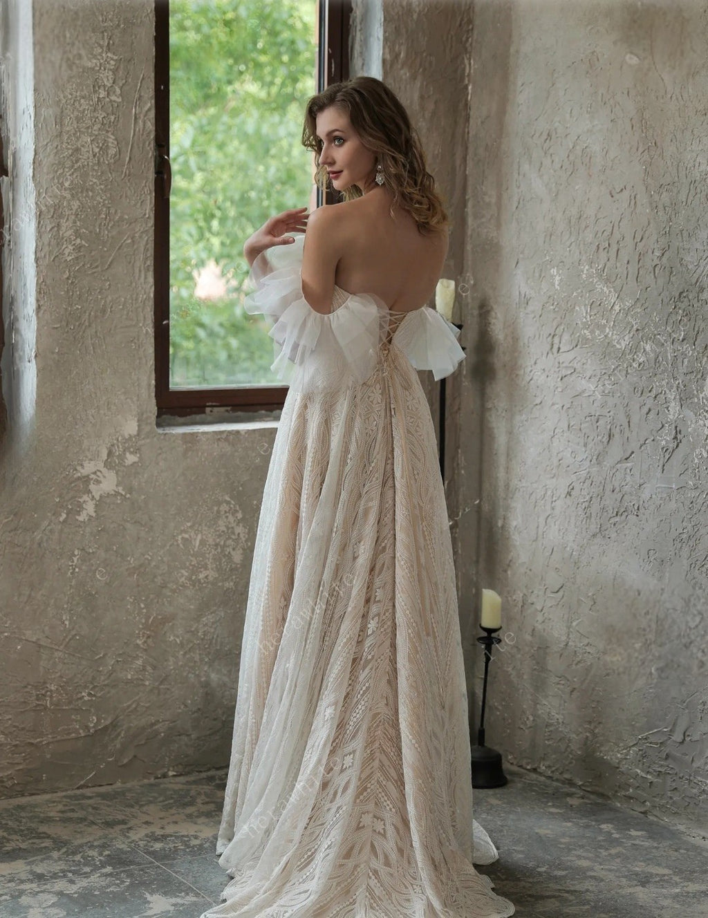 Woman in a white lace dress standing in a room with textured walls and a window.