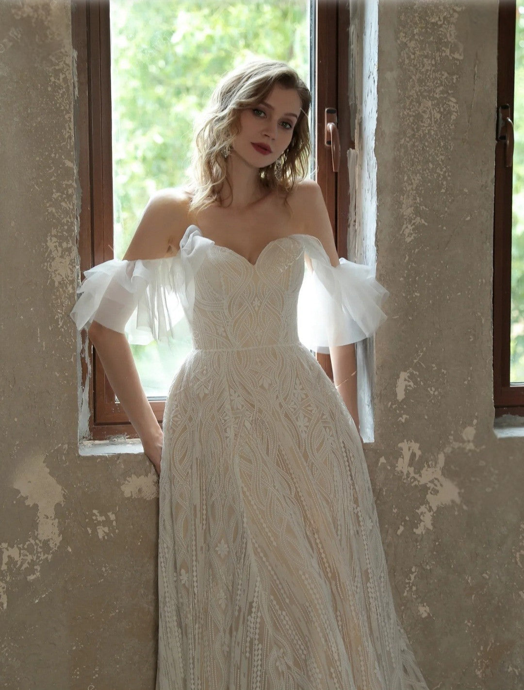 Woman in a white lace wedding dress standing in a room with peeling paint.