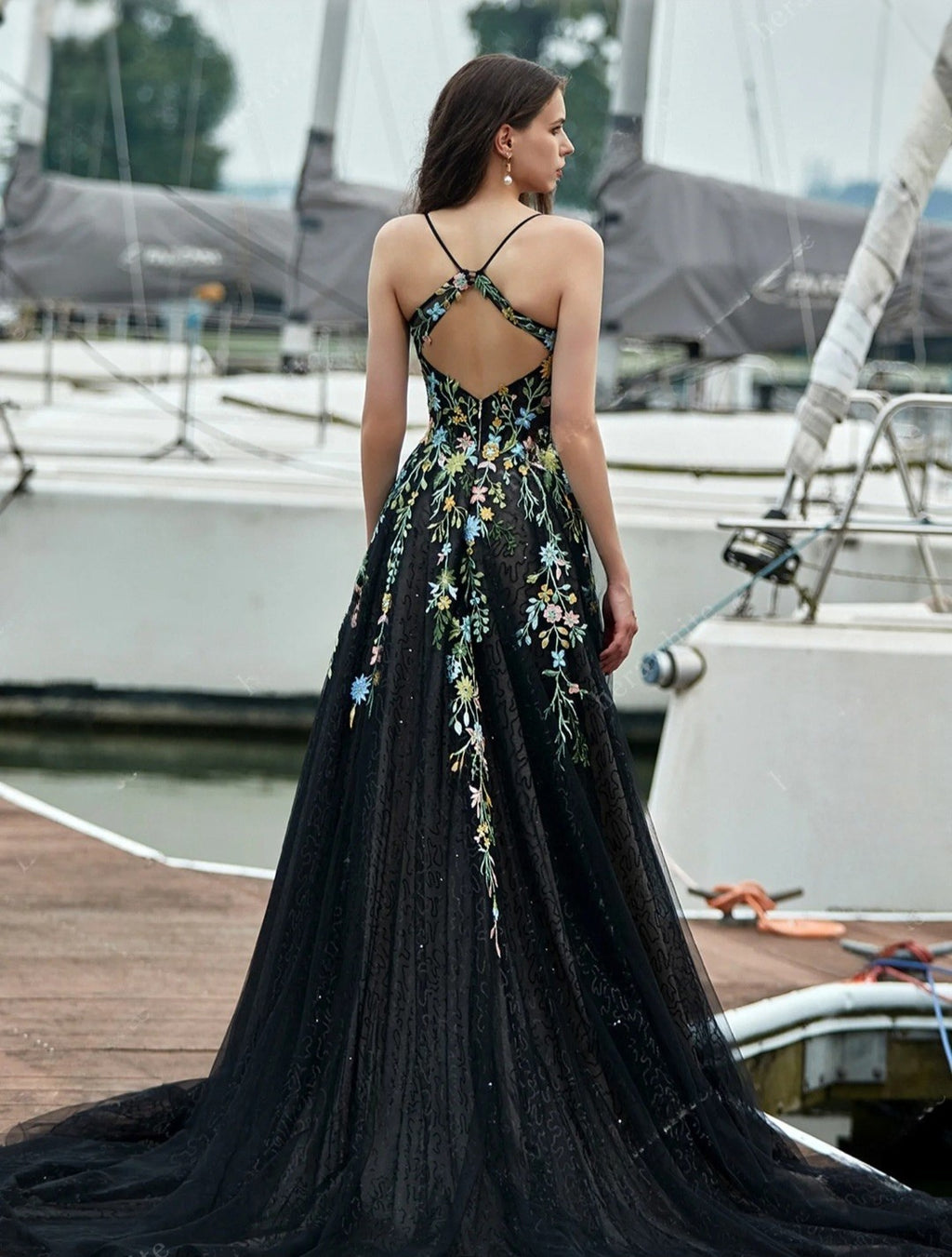 Woman in a black evening gown with floral embroidery standing on a dock with boats in the background