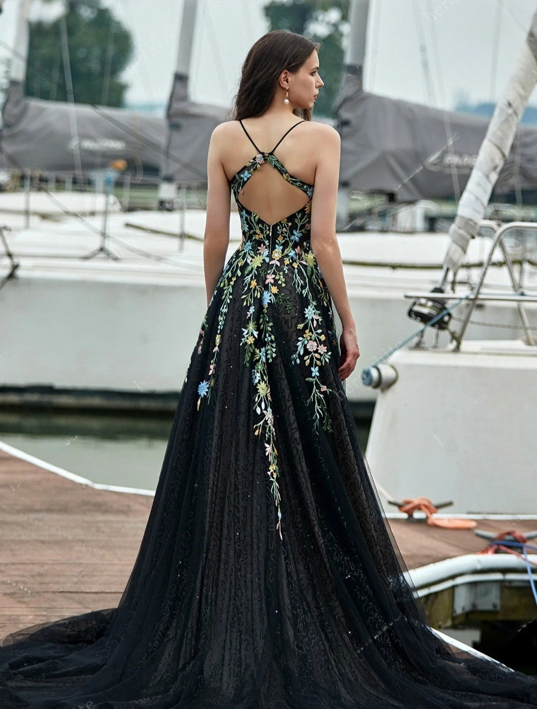 Woman in a black evening gown with floral embroidery standing on a dock with boats in the background