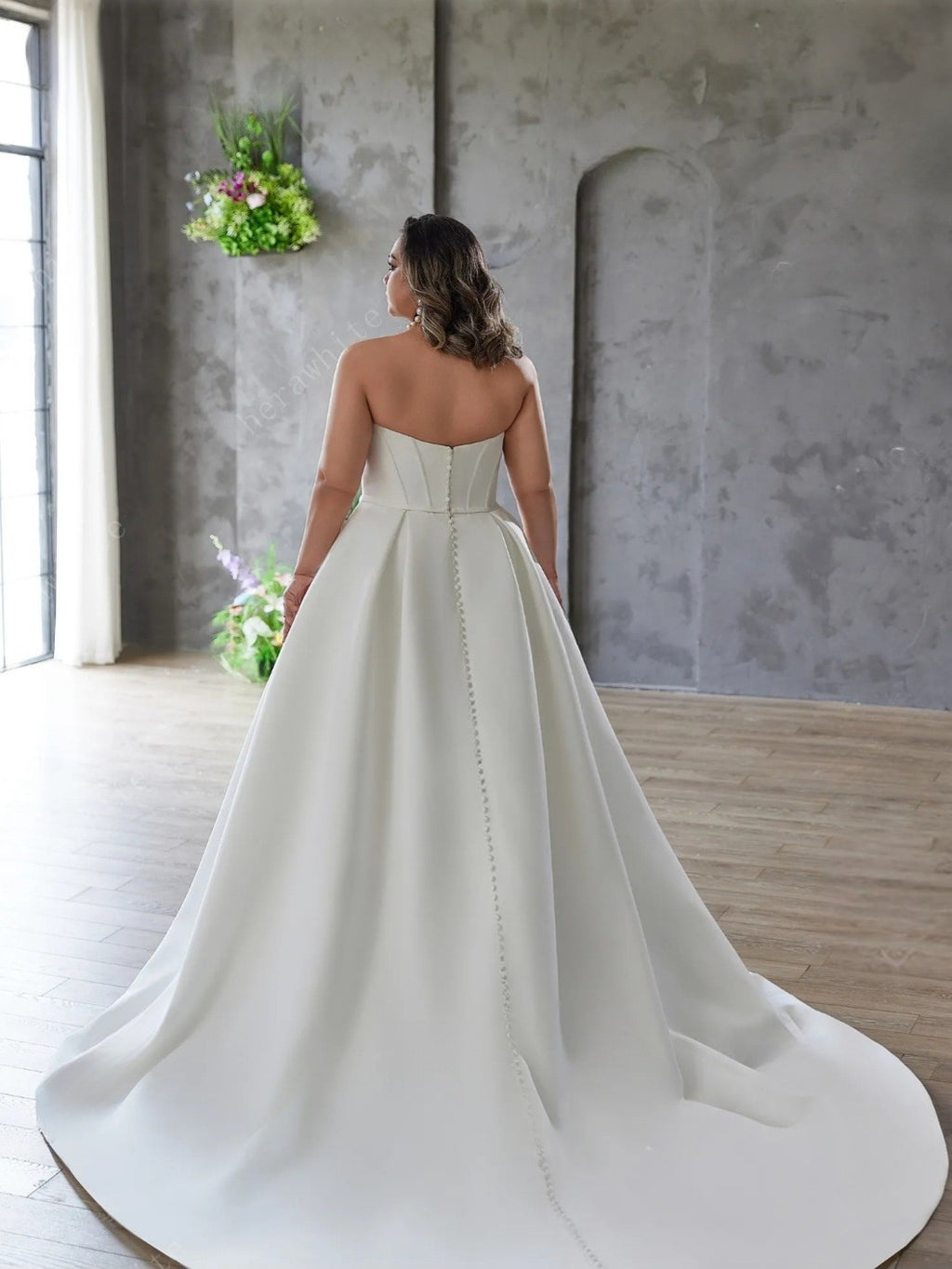 Woman in a strapless wedding dress standing in a room with a gray wall and wooden floor.