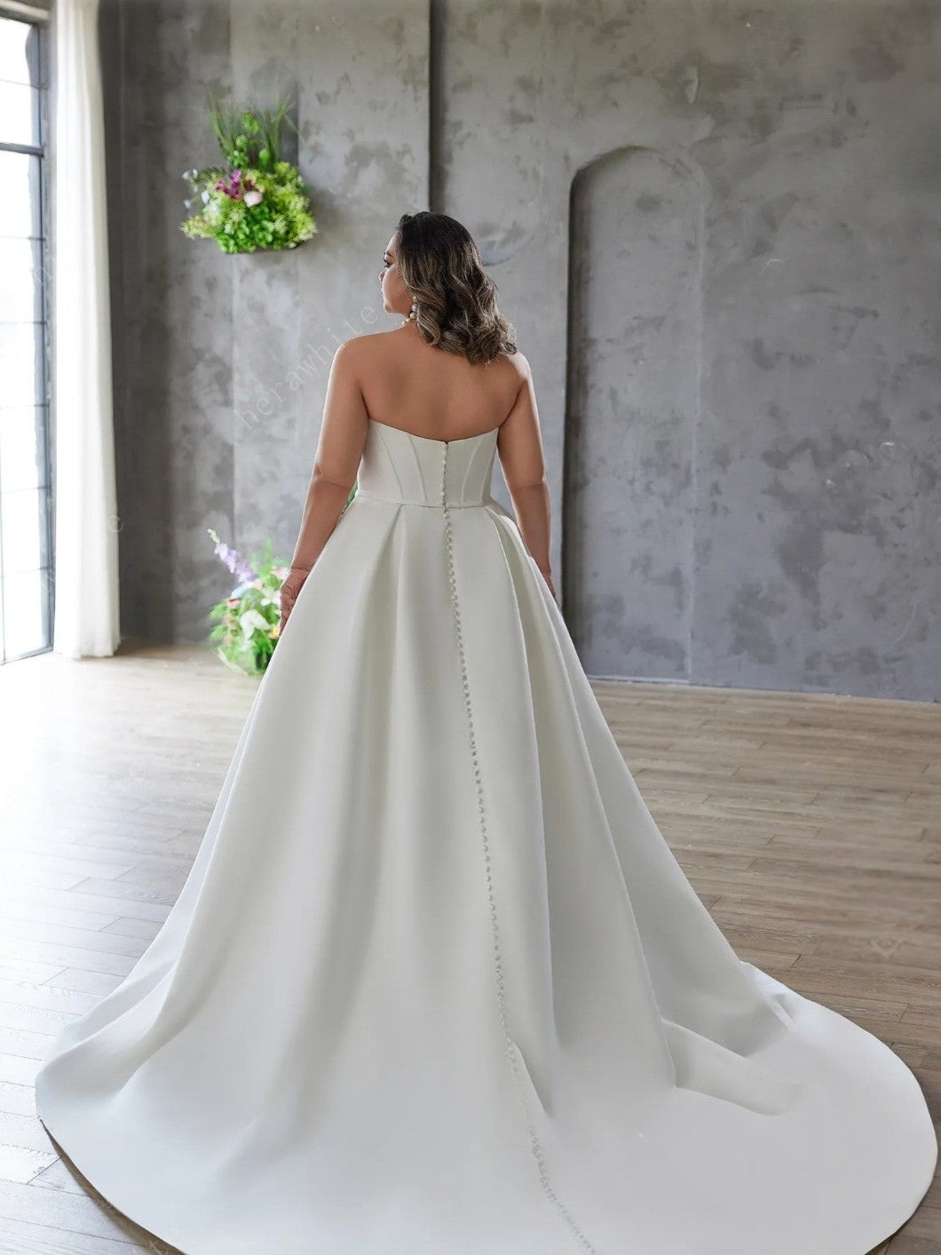 Woman in a strapless wedding dress standing in a room with a gray wall and wooden floor.
