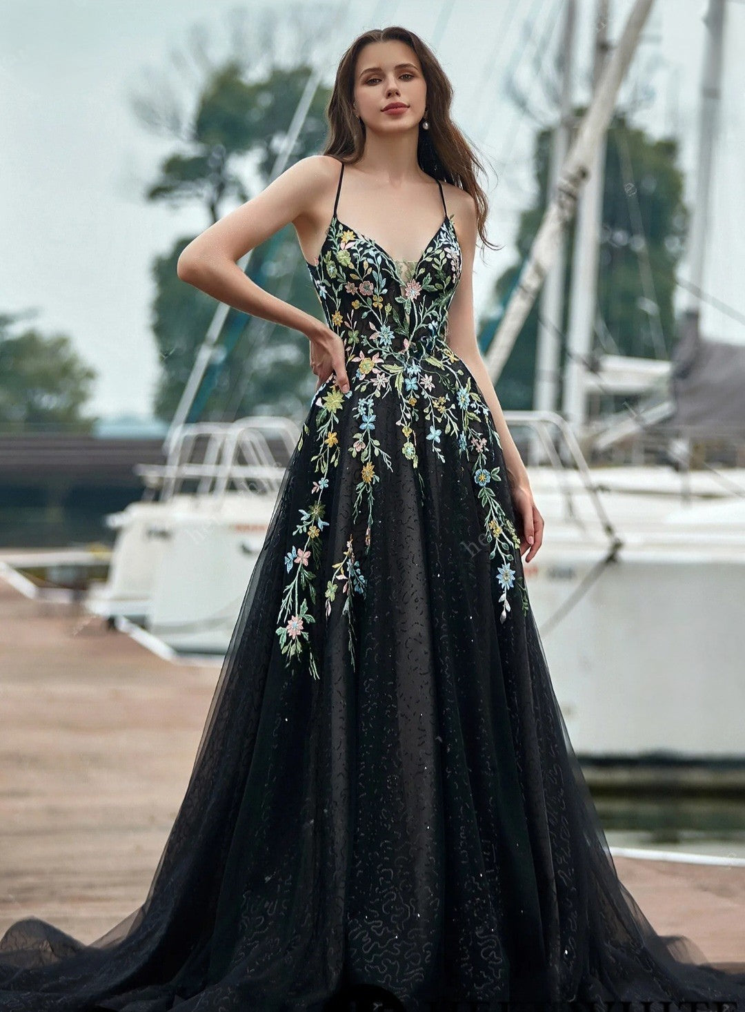 Woman in a black evening gown with floral embroidery standing on a dock with boats in the background.