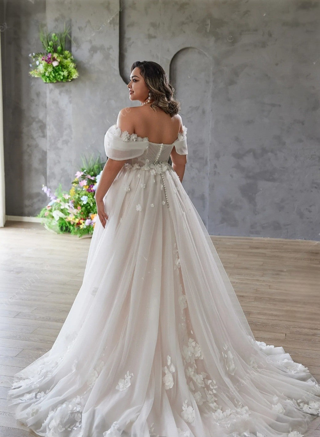 Woman in a white wedding dress standing in a room with flowers and a gray wall.