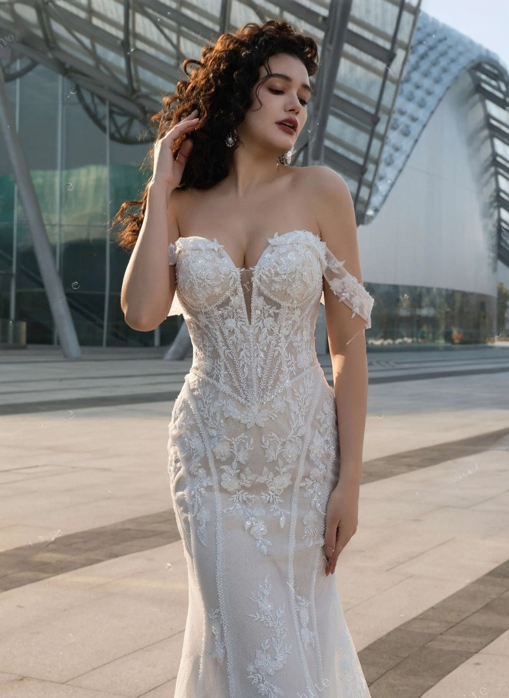 Woman in a white lace dress standing in front of modern architectural structures.