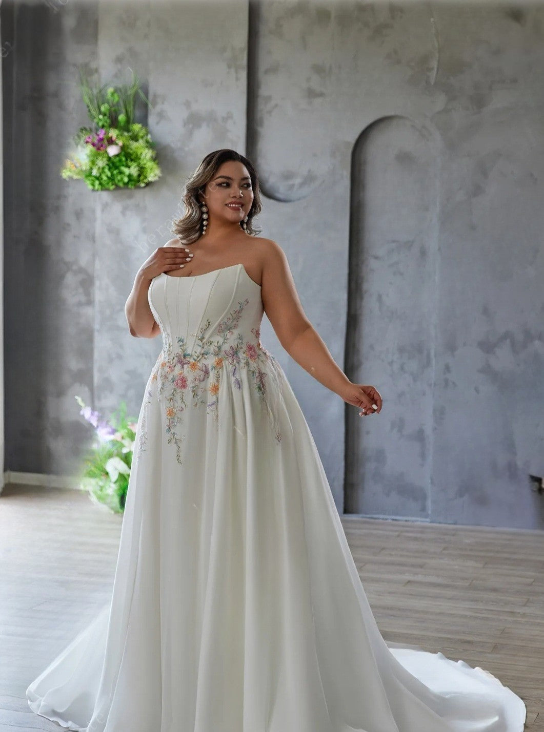 Woman in a white strapless gown with floral embroidery standing in a modern indoor setting.