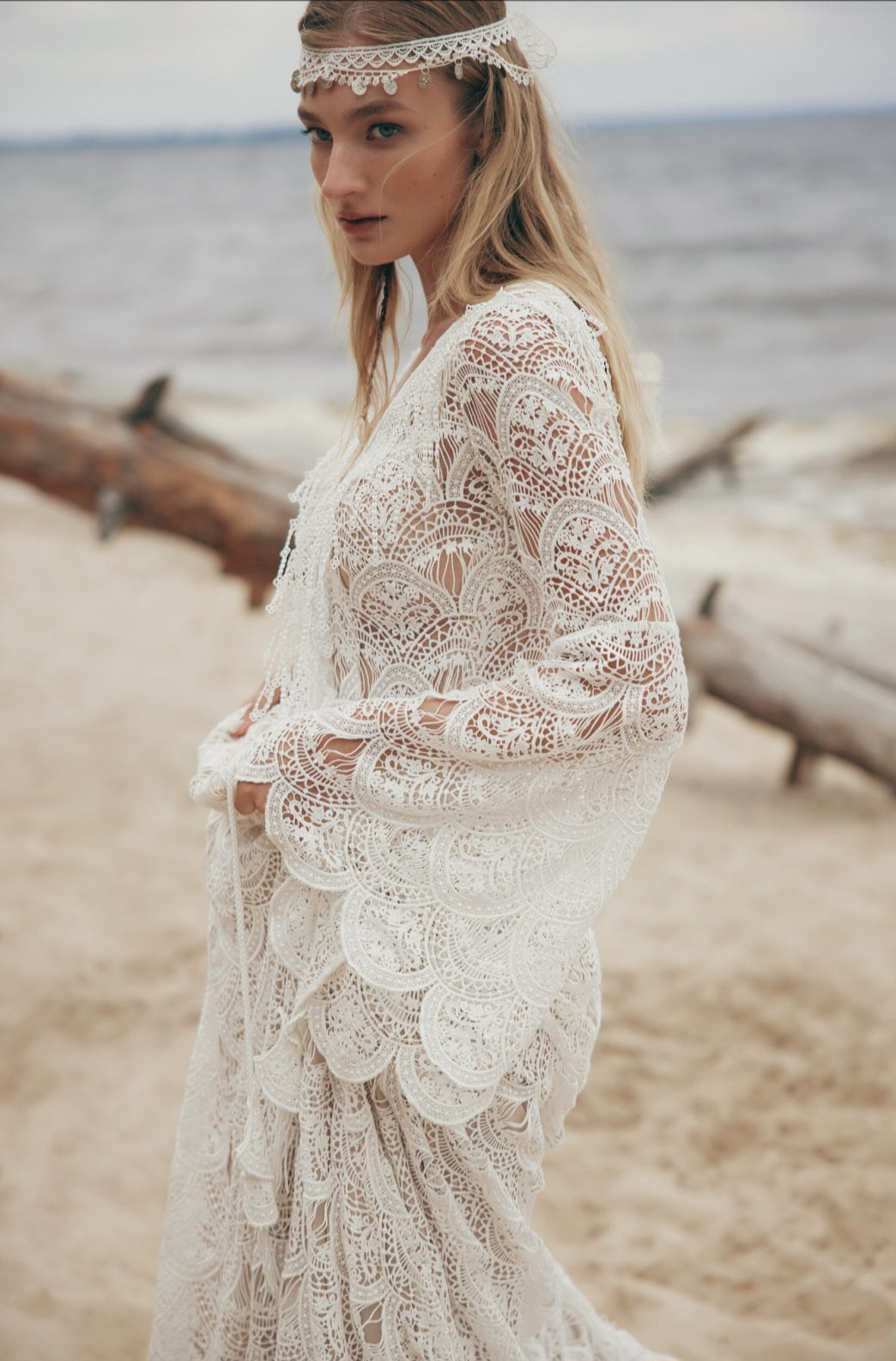 Woman in a lace dress standing on a beach with driftwood and water in the background