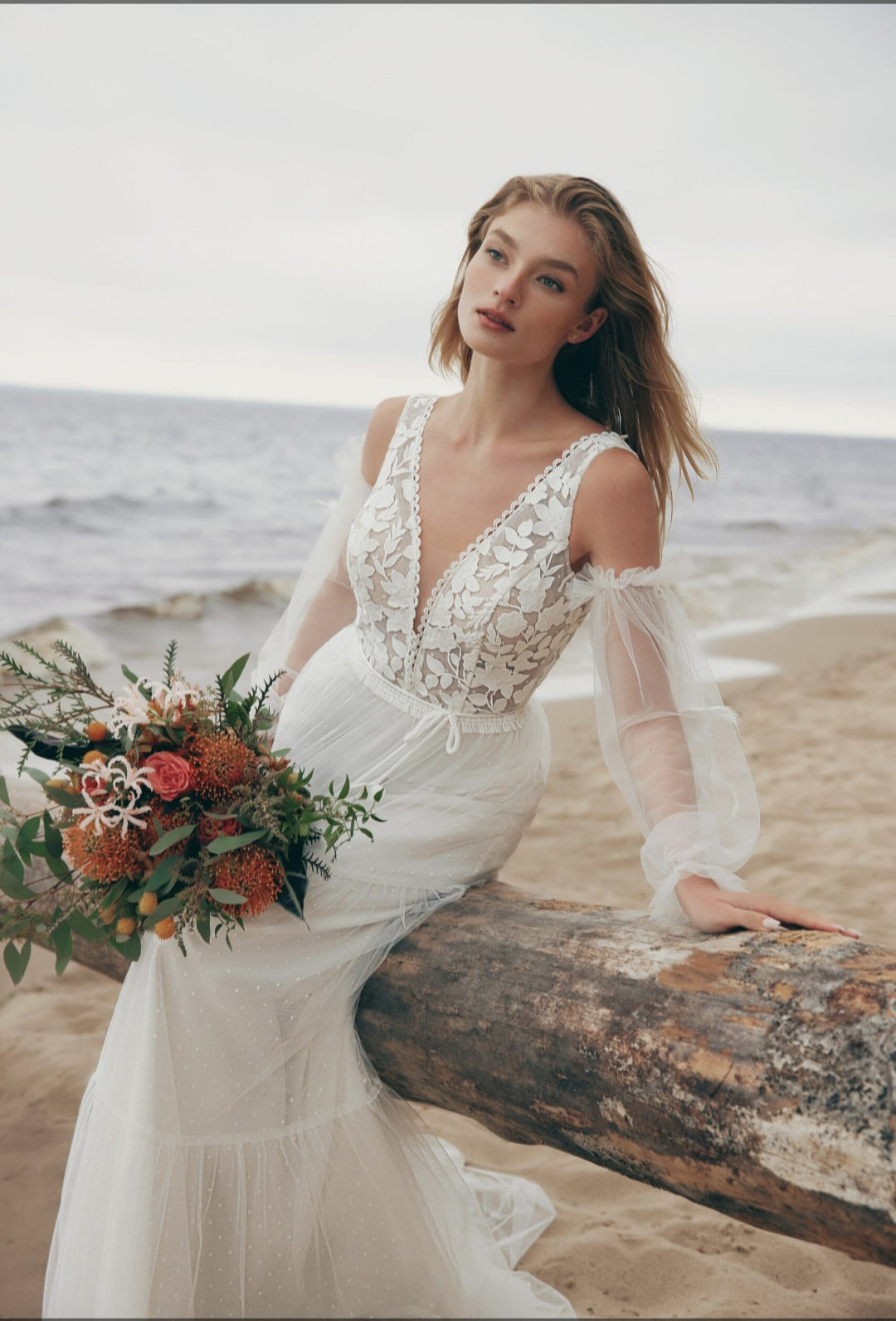 Woman in a white lace wedding dress holding a bouquet on a beach.