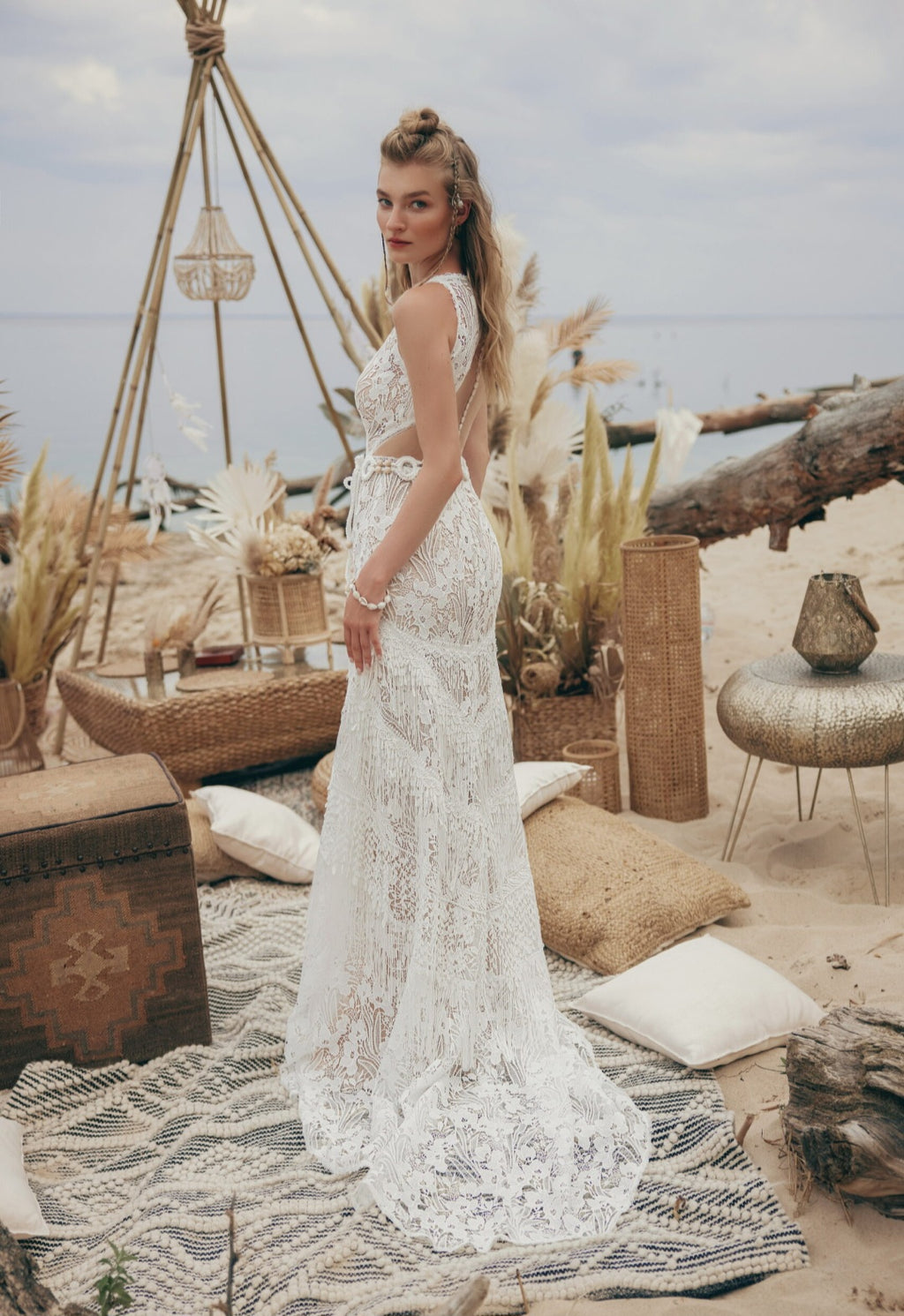 Woman in a white lace dress standing in a beachside setting with natural elements and tables.