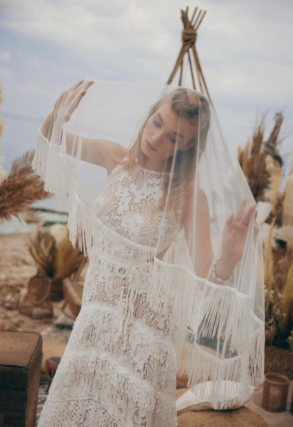 Woman in a white lace dress with fringe holding a veil against a natural background