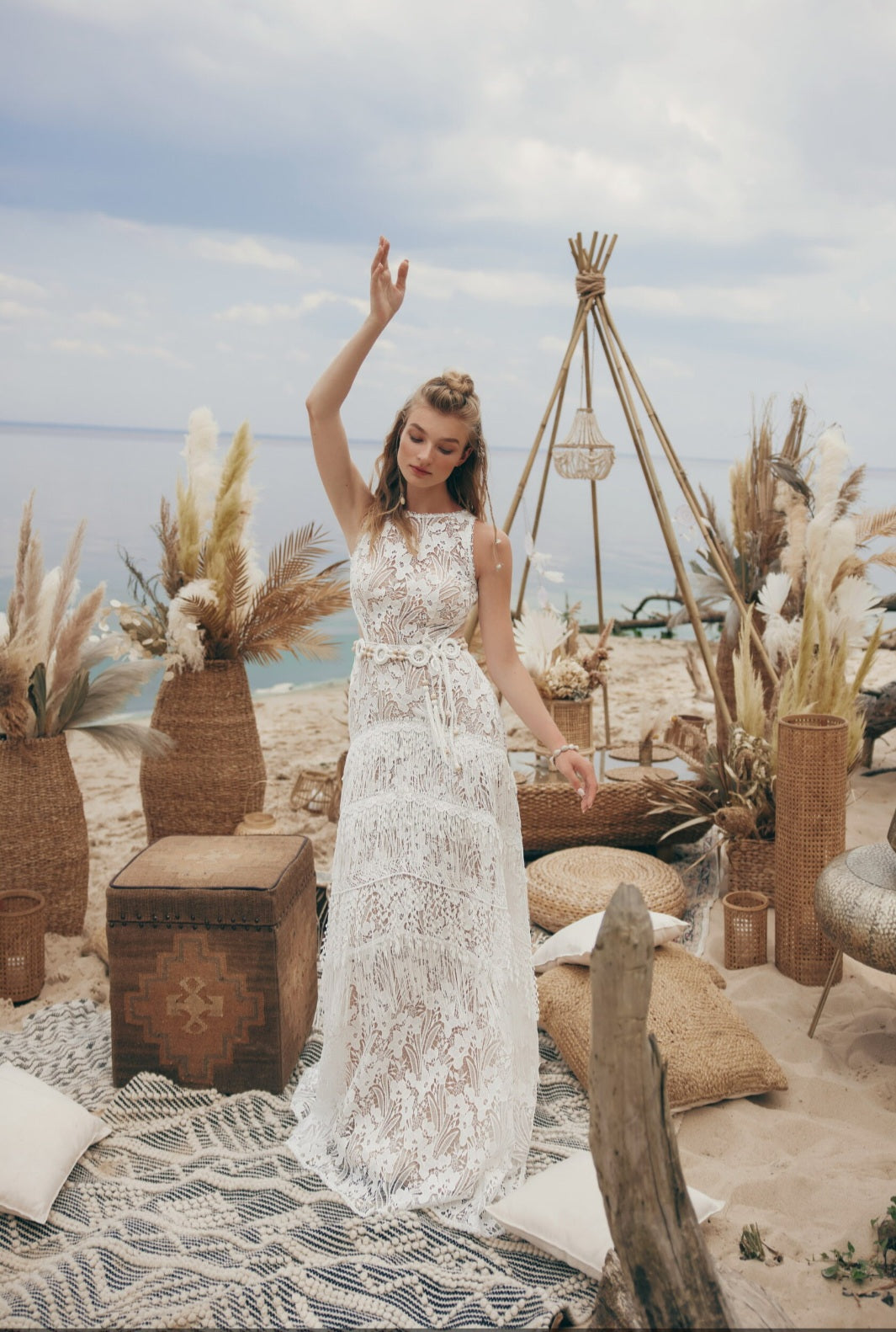 Woman in a white lace dress standing in a beach setting with decorative plants and furniture.