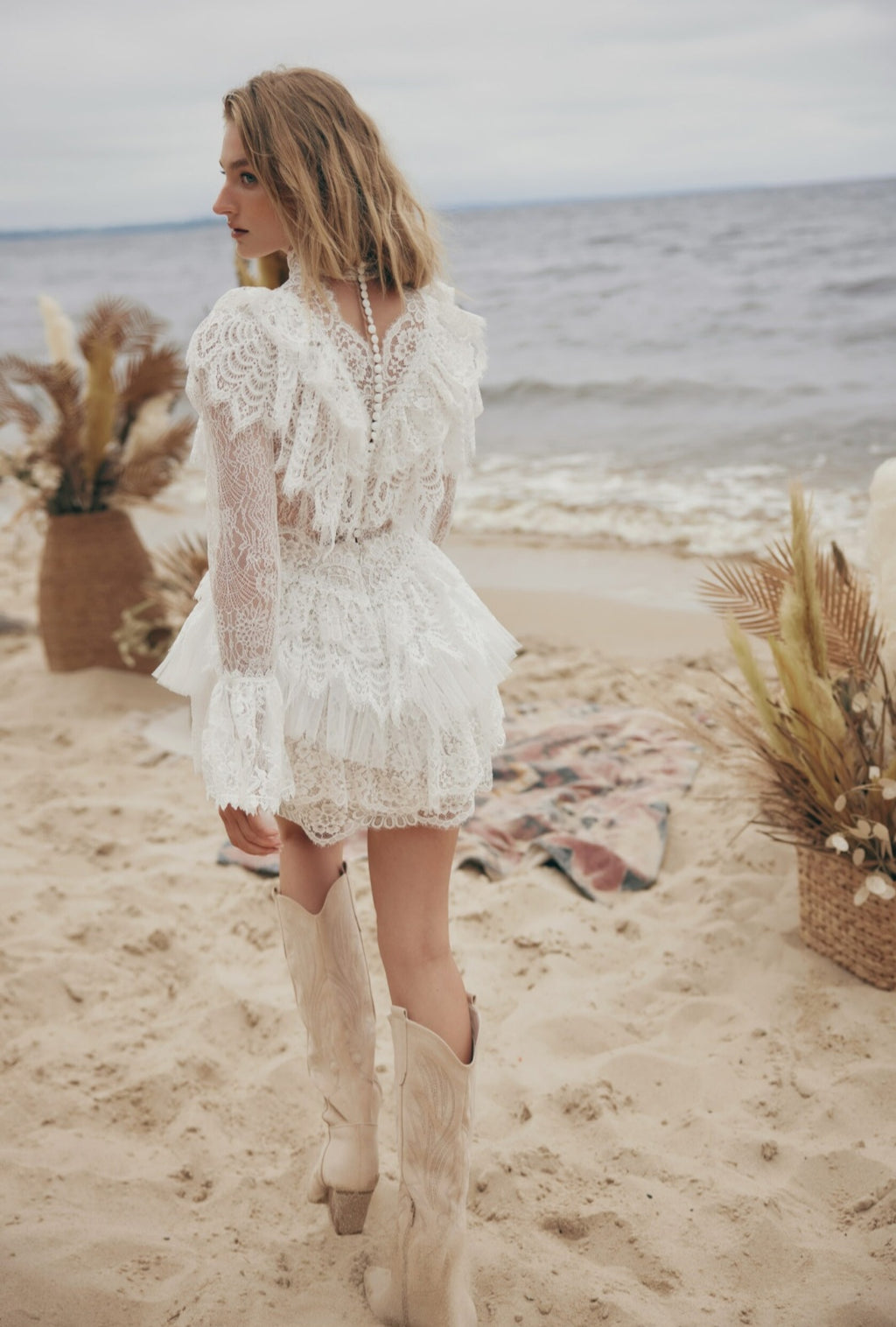Woman in a white lace dress and boots standing on a beach with decorative elements.