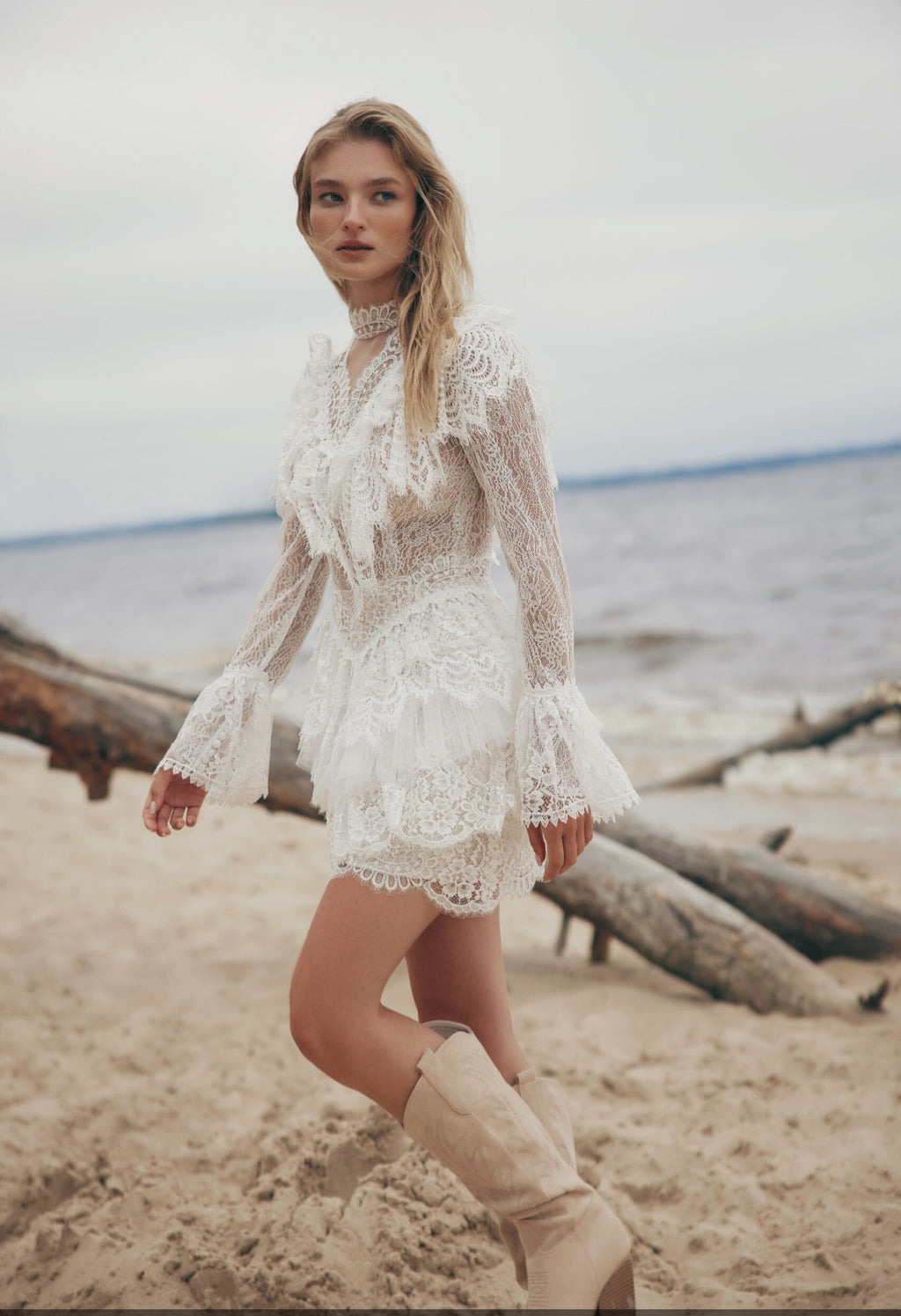Woman in a white lace dress standing on a beach with logs in the background
