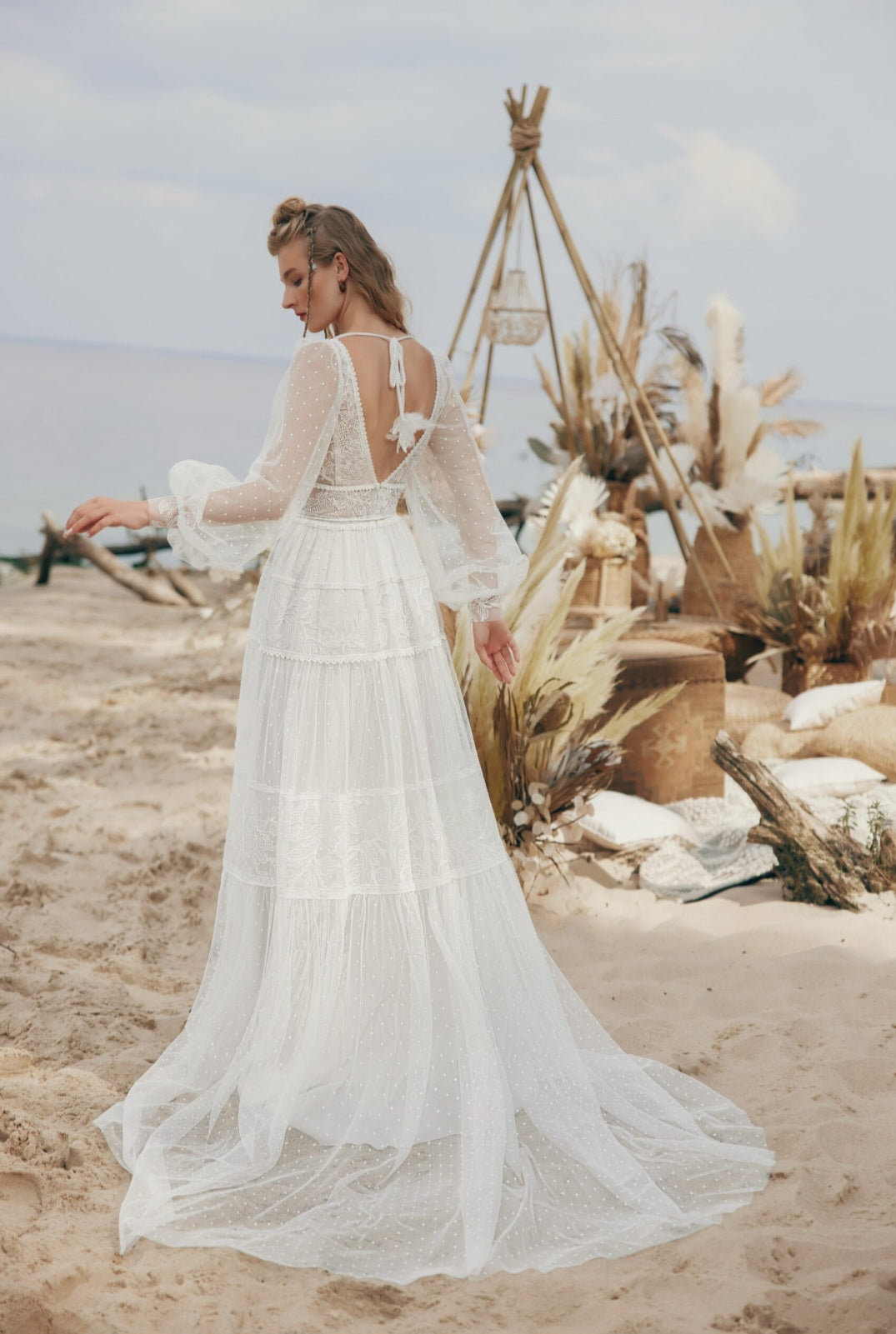 Woman in a white bohemian-style dress standing on a sandy beach with desert plants and a teepee-like structure.