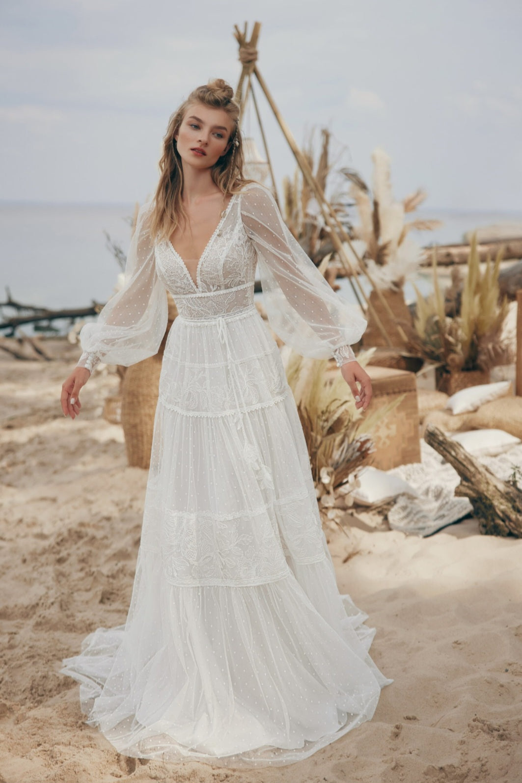 Woman in a white bohemian-style dress standing on a sandy beach with natural elements in the background.