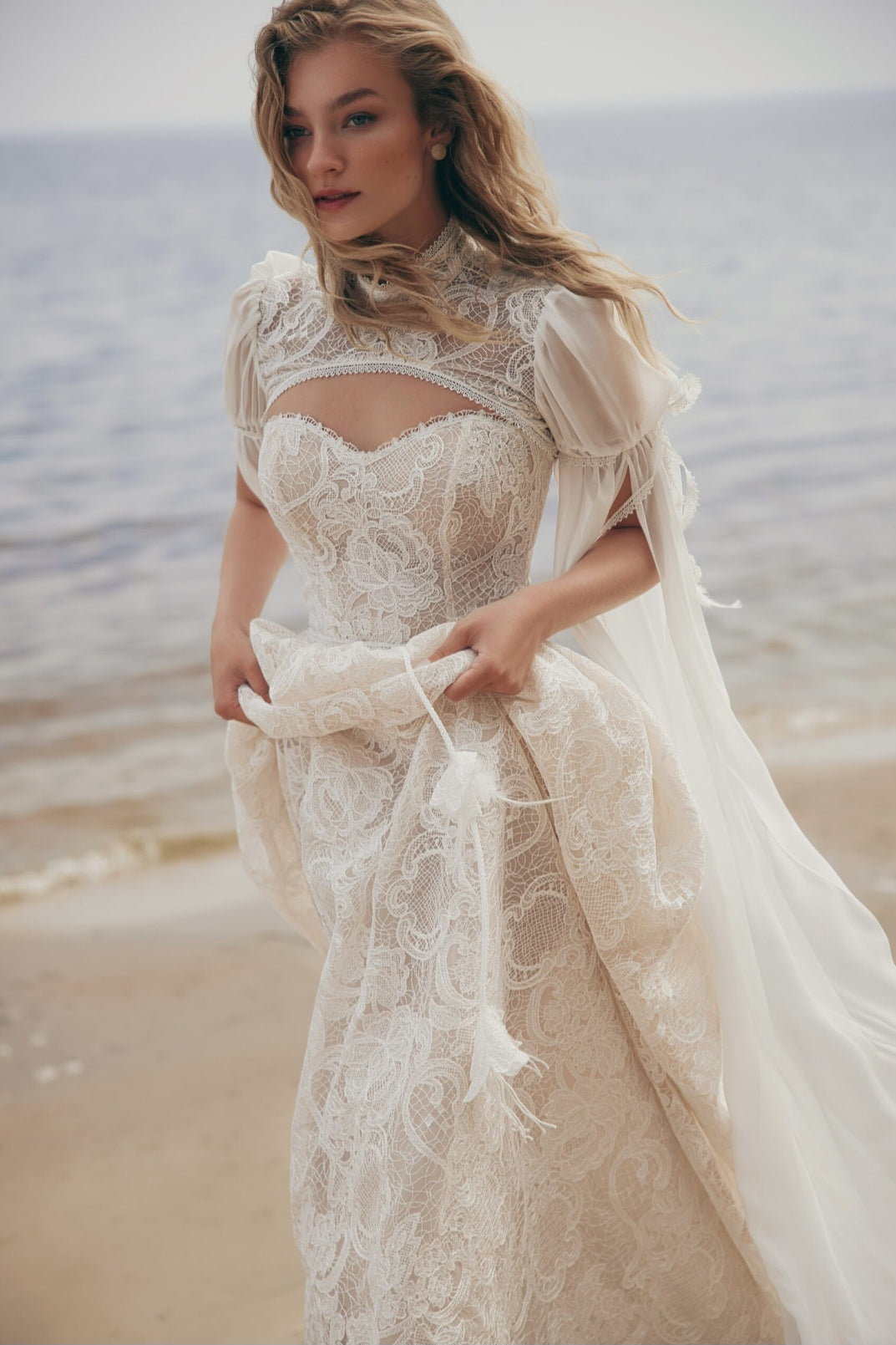 Woman in a lace wedding dress standing on a beach.