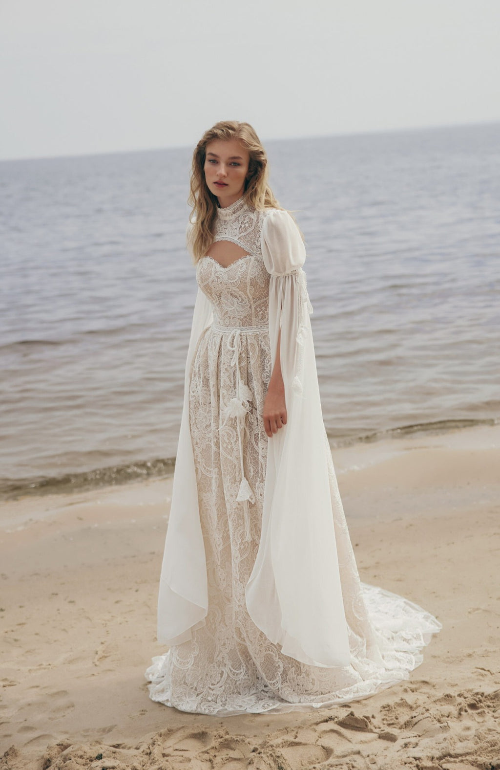 Woman in a lace wedding dress standing on a beach with ocean in the background