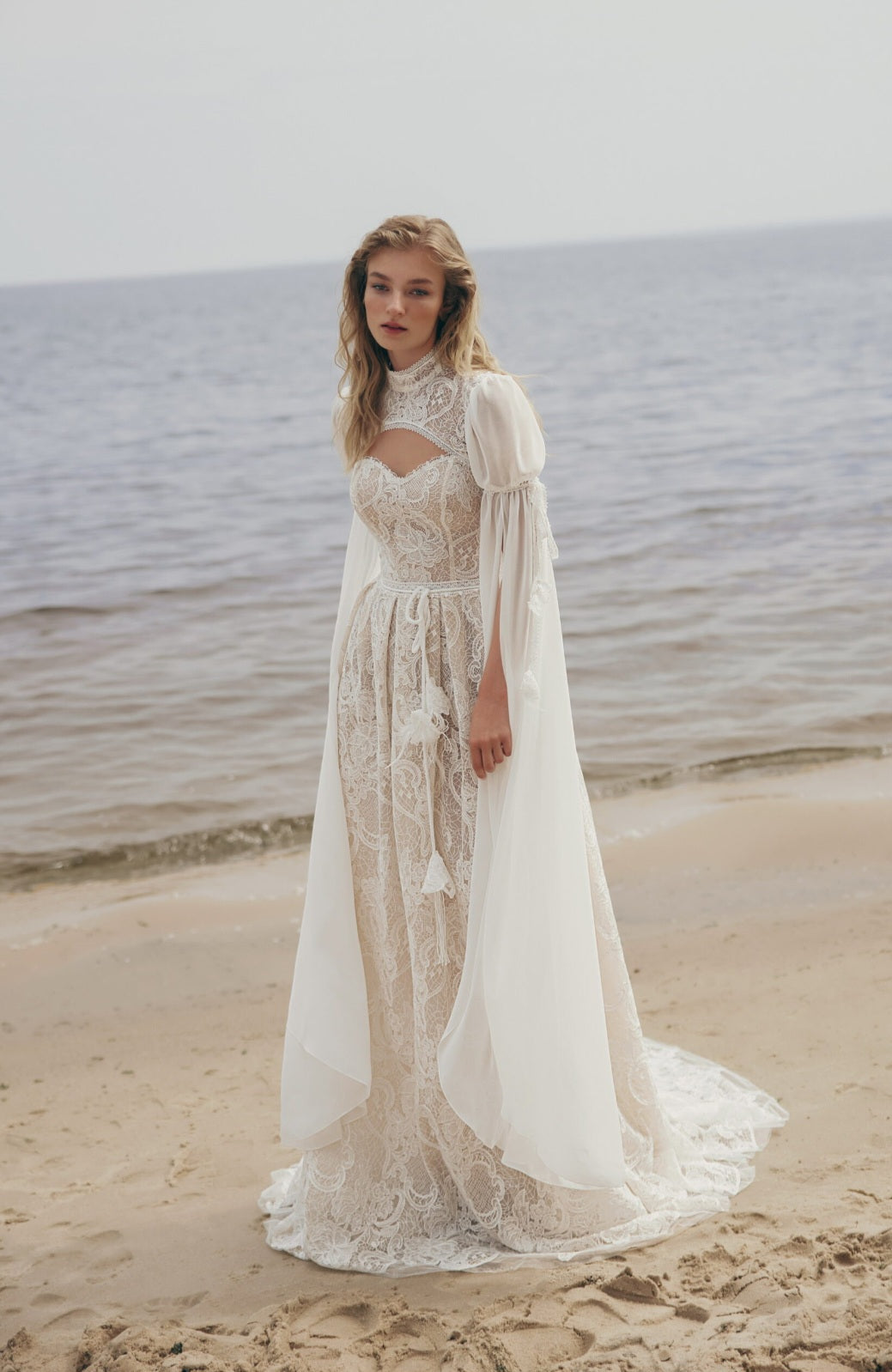 Woman in a lace wedding dress standing on a beach with ocean in the background