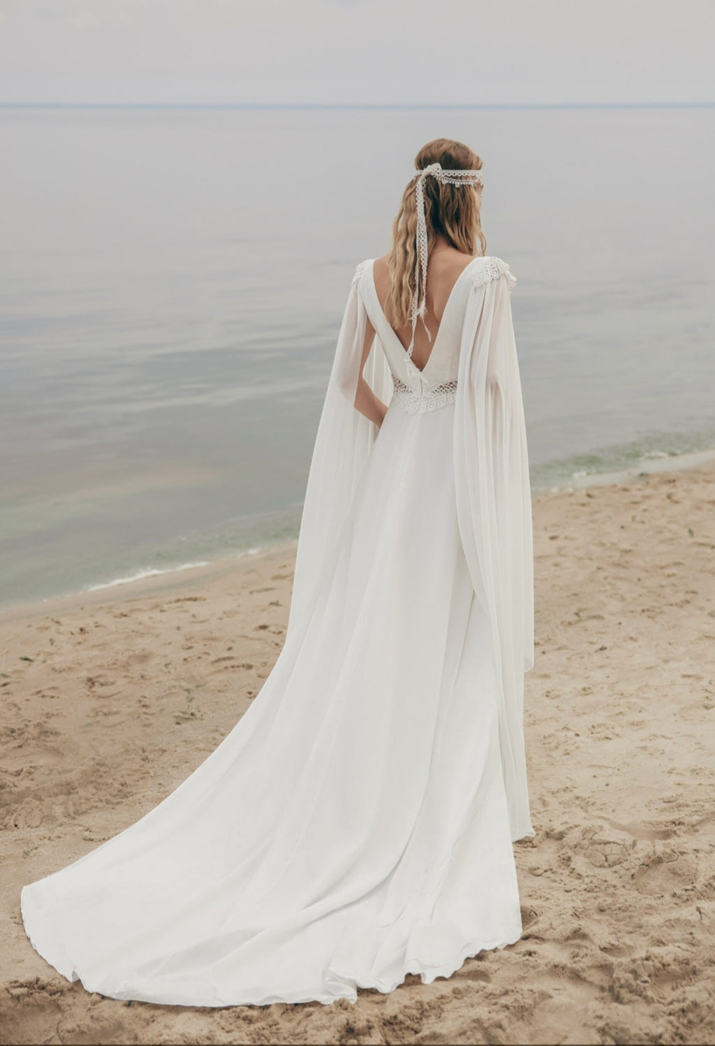 Woman in a white wedding dress with a cape standing on a beach.