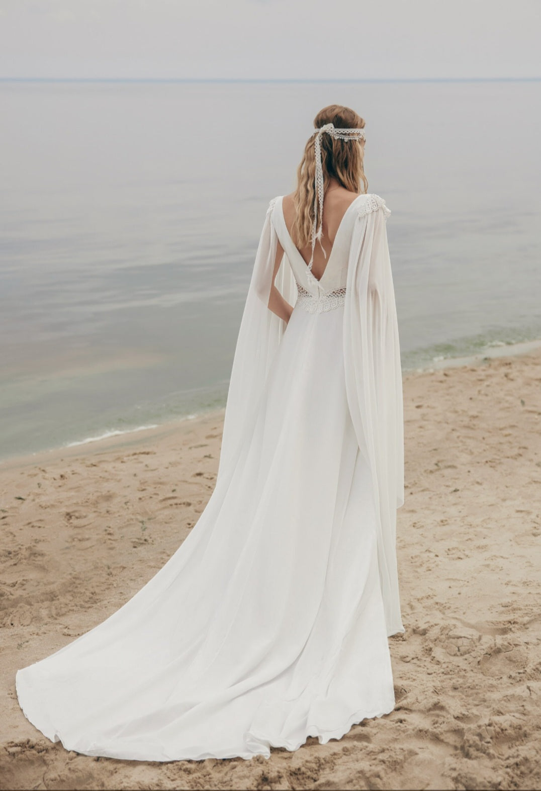 Woman in a white wedding dress with a cape standing on a beach.