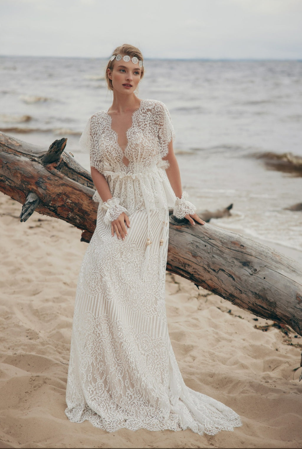 Woman in a lace wedding dress standing on a beach with driftwood.