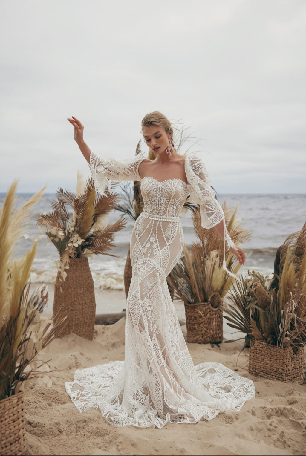 Woman in a lace wedding dress standing on a beach with decorative plants and ocean view.