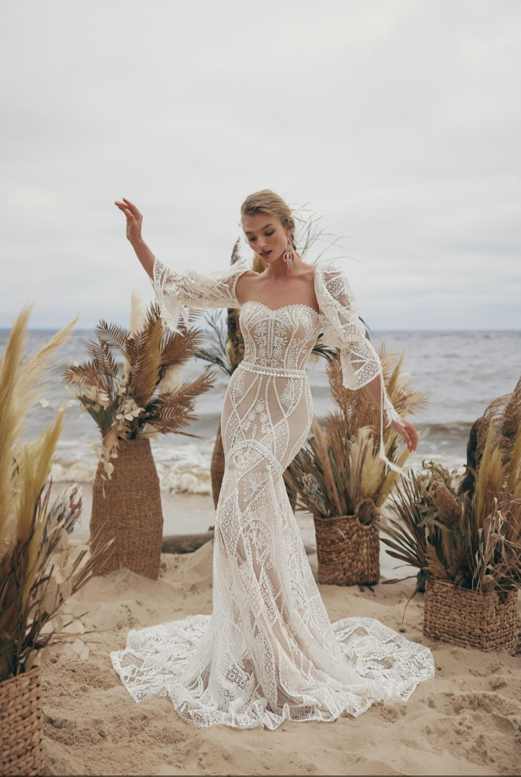 Woman in a lace wedding dress standing on a beach with decorative plants and ocean view.