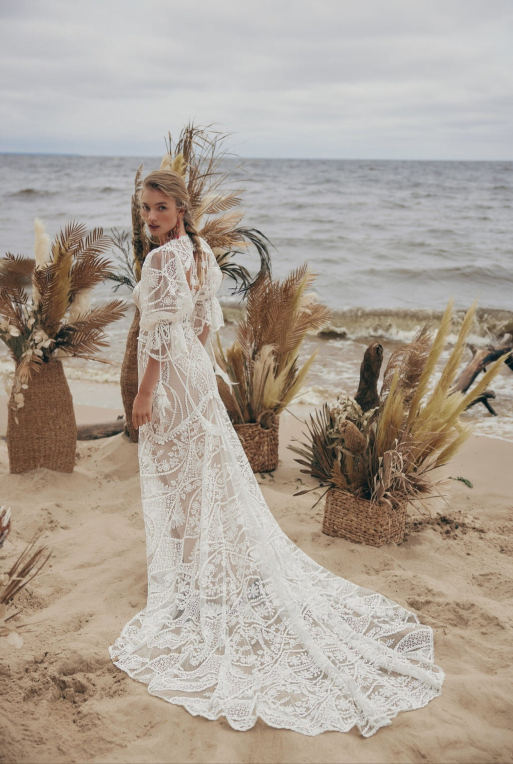 Woman in a lace wedding dress standing on a beach with decorative plants.