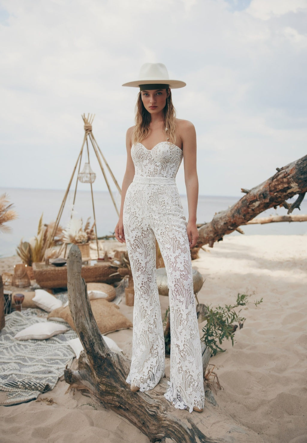 Woman in a white lace jumpsuit and hat standing on a beach with driftwood and a teepee in the background.