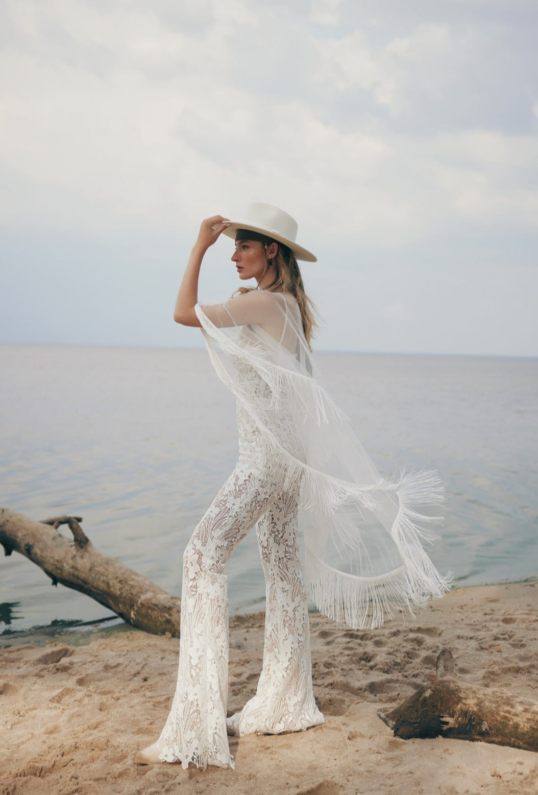 Woman in a white lace outfit and hat standing on a beach with ocean in the background