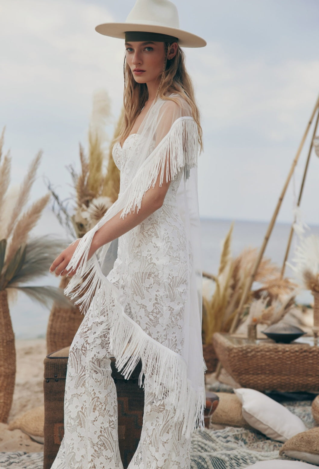 Woman in a white fringe dress and hat standing in a desert-like setting with desert plants and a bowl.