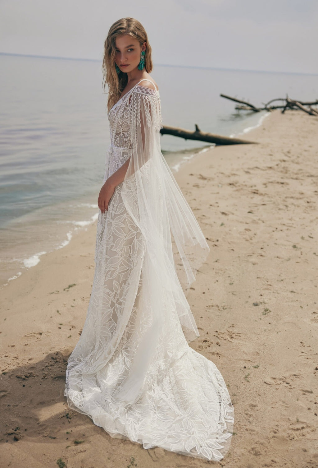 Woman in a white lace dress standing on a beach with water and driftwood in the background.