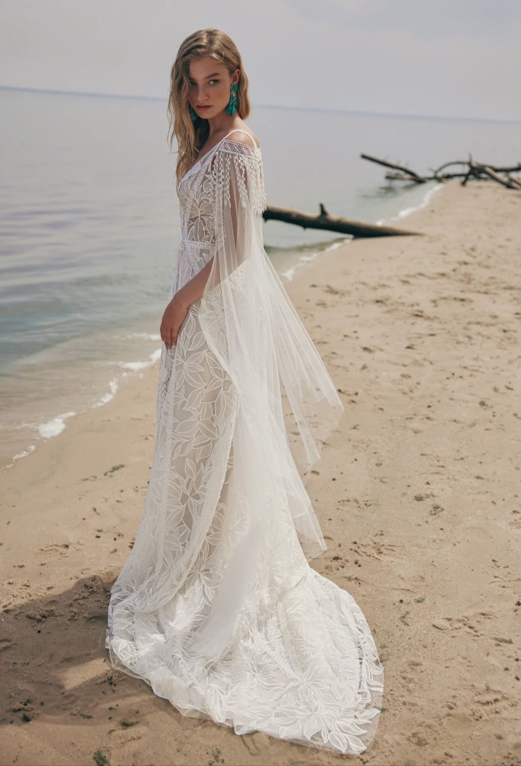 Woman in a white lace dress standing on a beach with water and driftwood in the background.