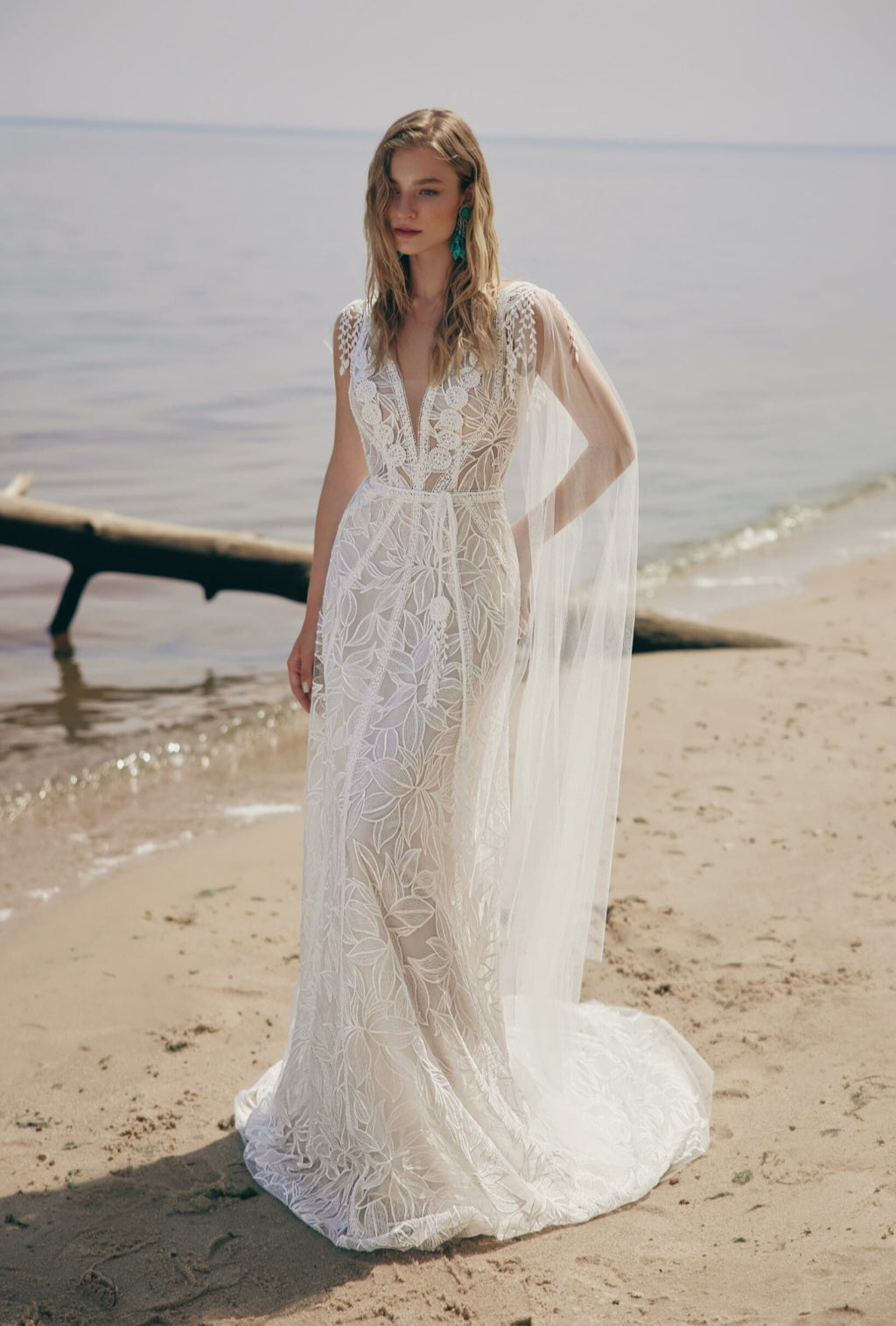 Woman in a lace wedding dress standing on a beach with water in the background