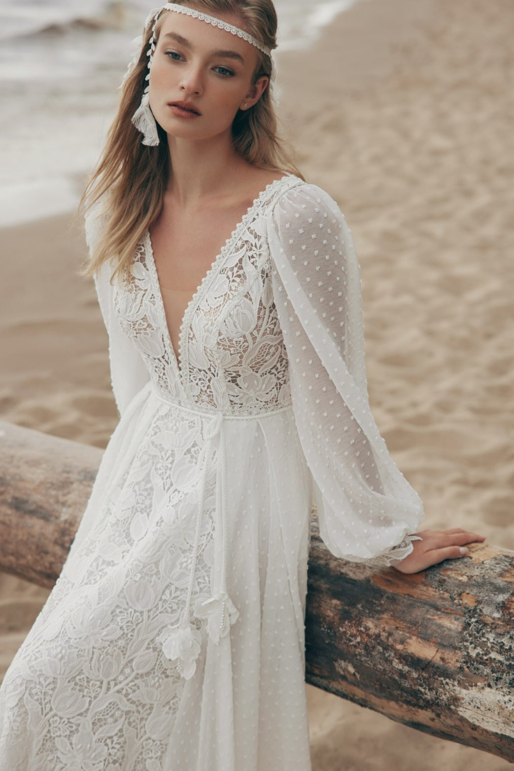 Woman in a white lace dress standing on a beach with sand and water in the background.