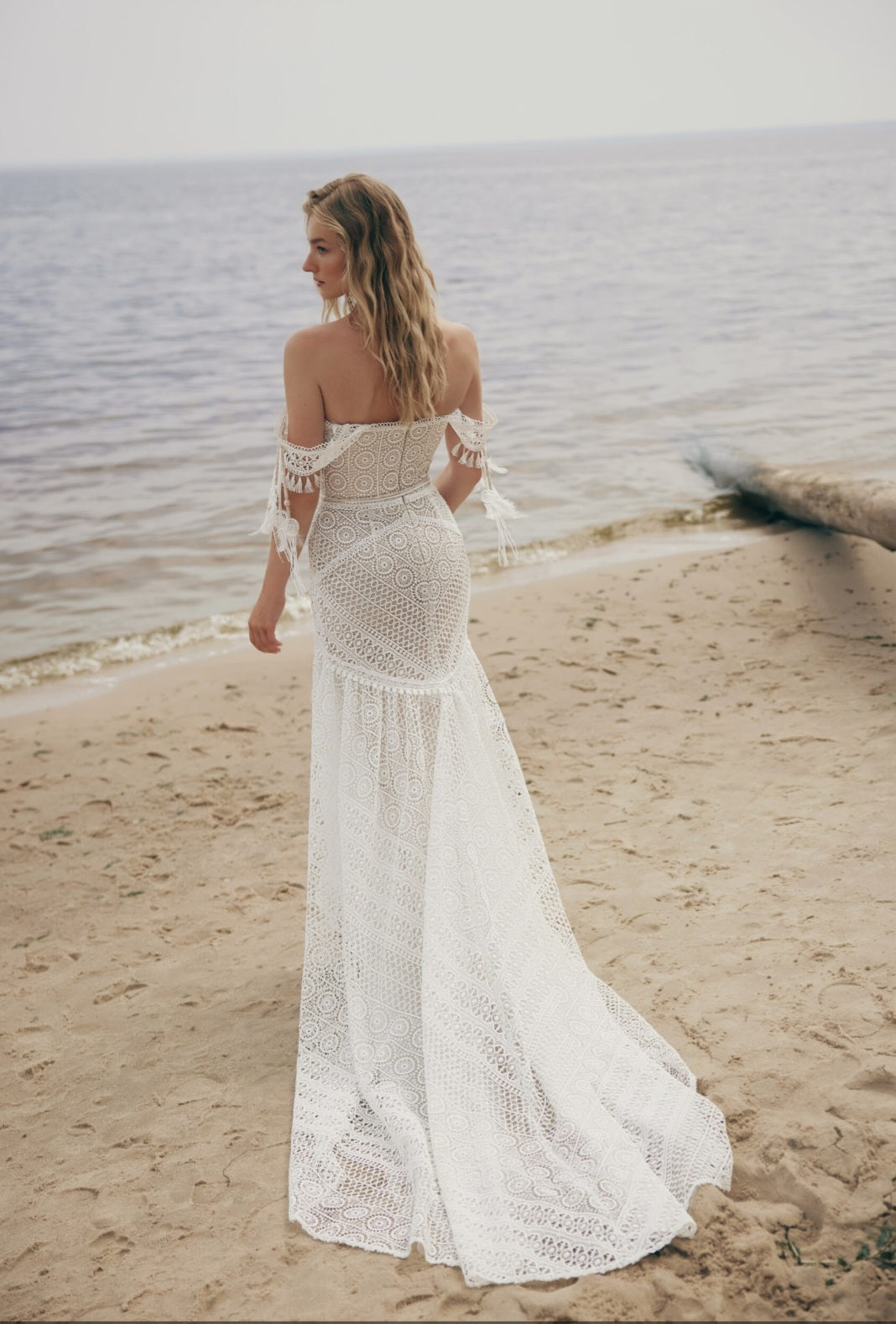 Woman in a white lace dress standing on a beach.