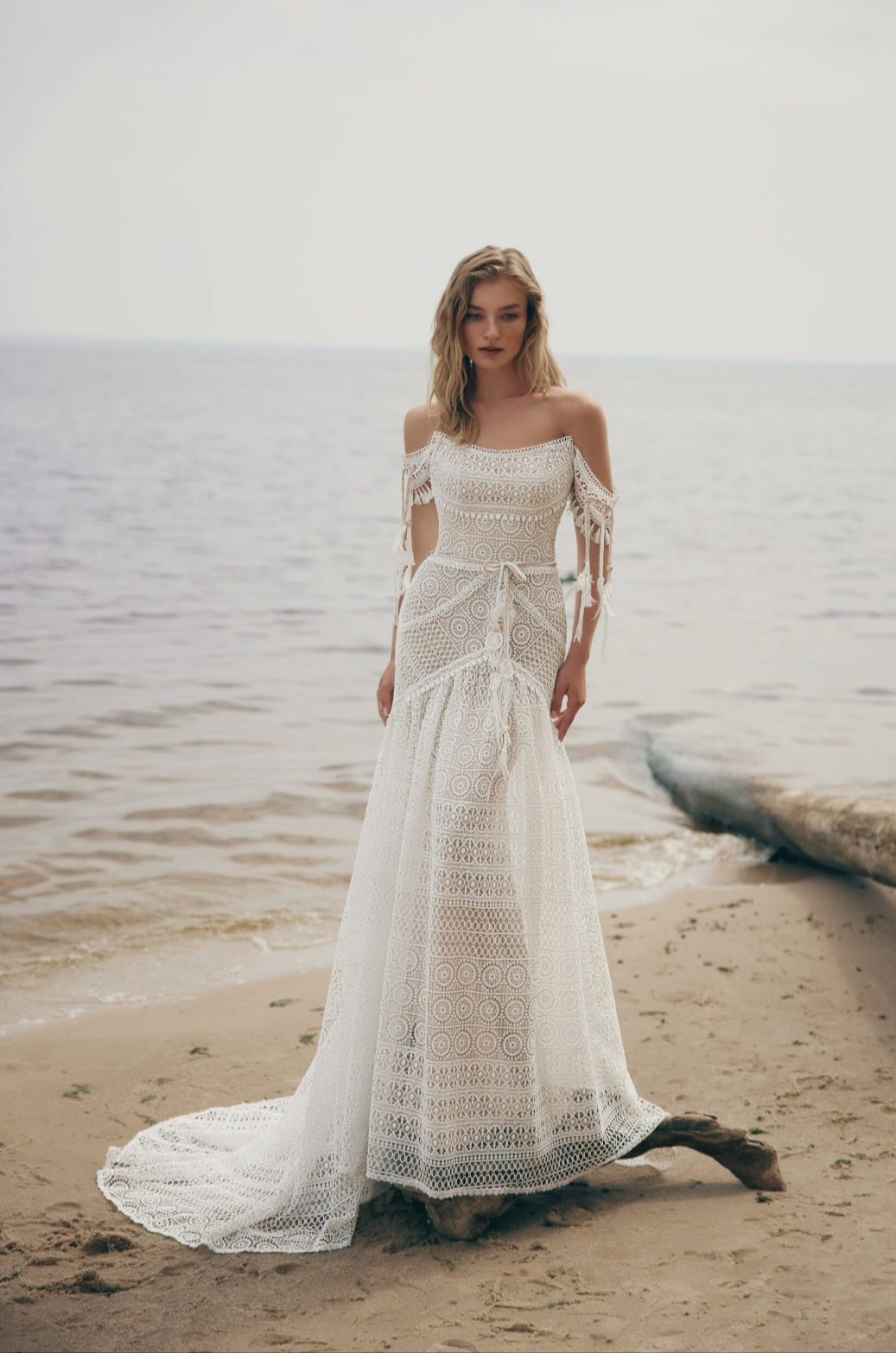 Woman in a white lace dress standing on a beach.