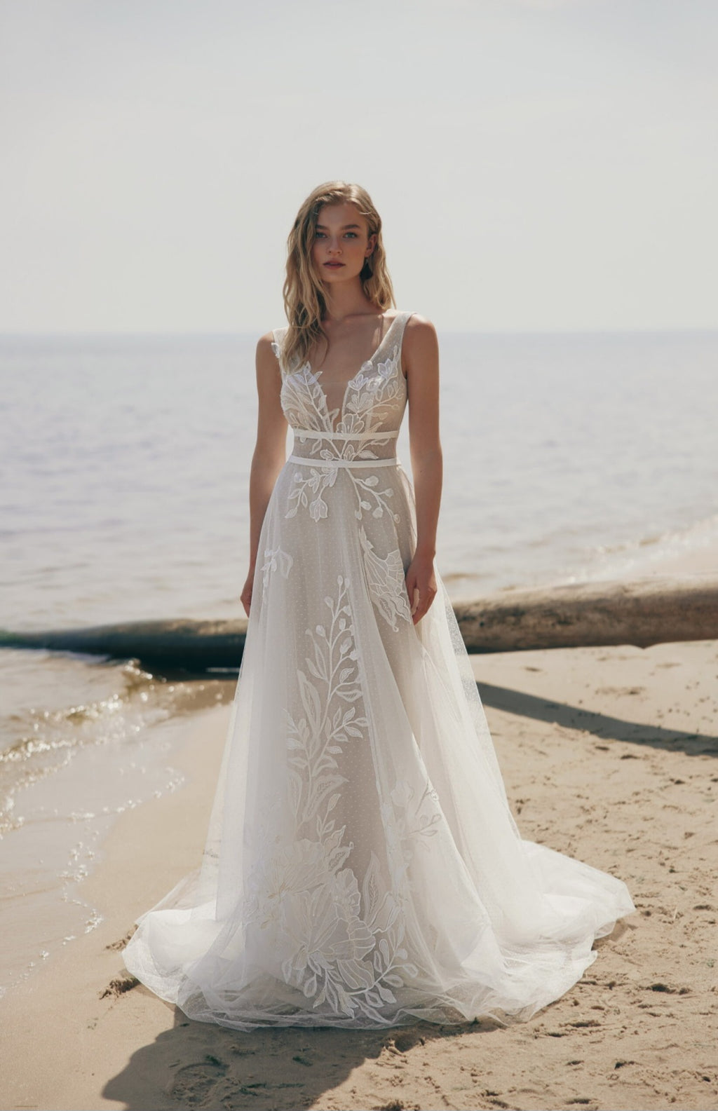 Woman in a white lace wedding dress standing on a beach.