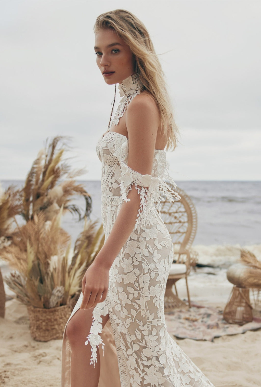 Woman in a white lace dress on a beach with ocean and plants in the background