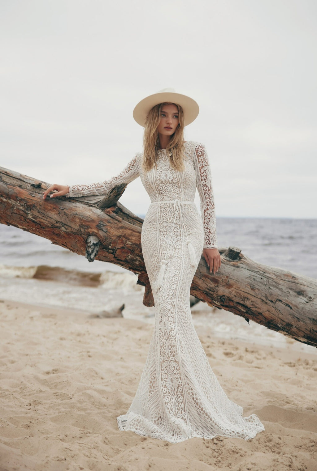 Woman in a lace dress and hat leaning against a log on a beach