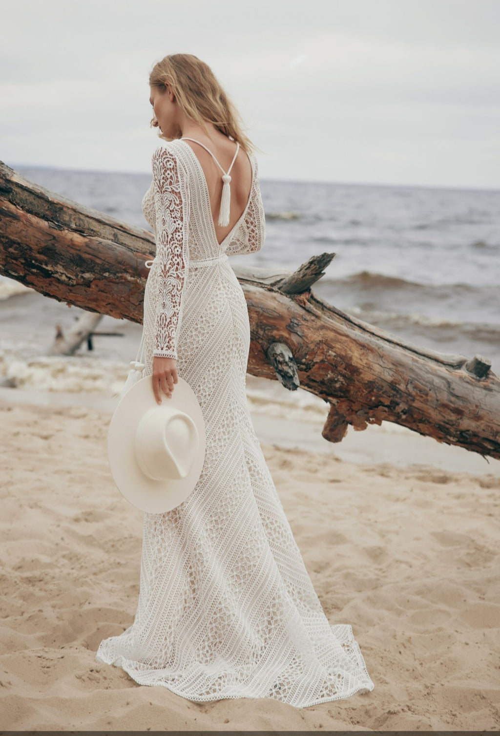 Woman in a white lace dress standing on a beach with a large log and ocean in the background